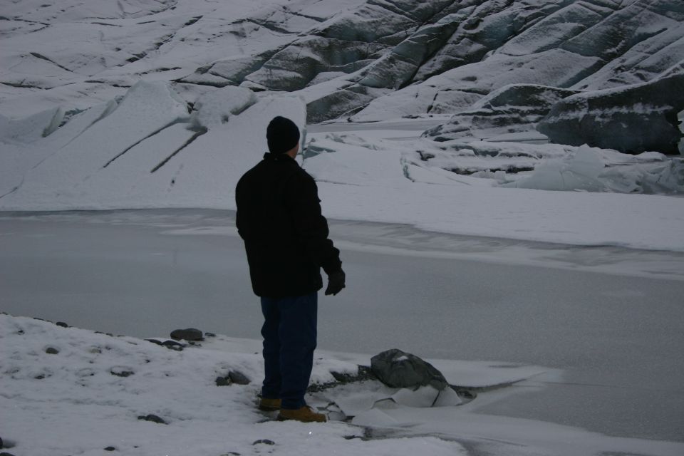 Person in black jacket and blue jeans stands on snowy ground looking at a frozen body of water with a glacier behind