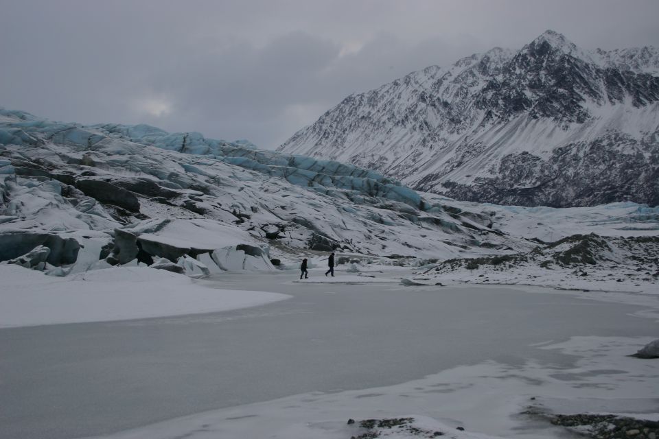 Snow-covered glacier and mountains under a cloudy sky with two people walking on ice