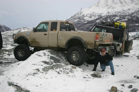 2003 Jeep TJ - Two off-road vehicles are parked in a snowy, mountainous landscape, with a person crouching near the rear of the pickup truck.