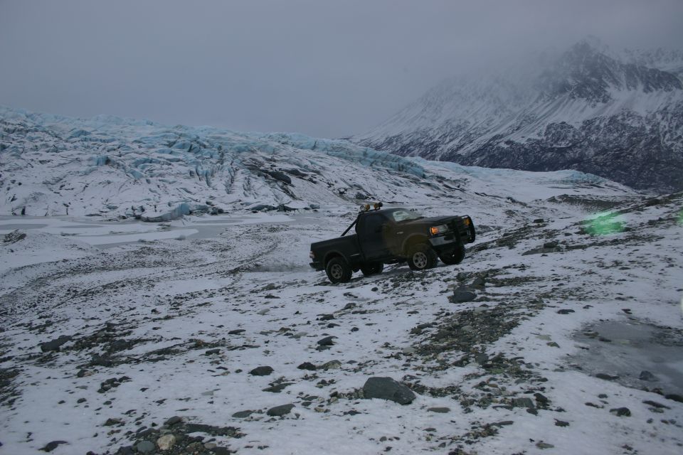 Pickup truck driving on a snowy, rocky slope with a glacier and mountains in the background