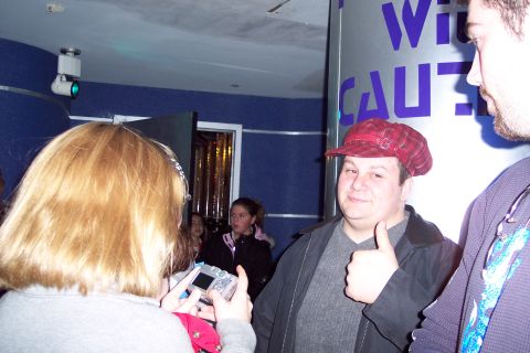 2001 Ford Crown Victoria - A man in a red plaid cap gives a thumbs-up gesture while standing in front of a large cylindrical sign with purple lettering.