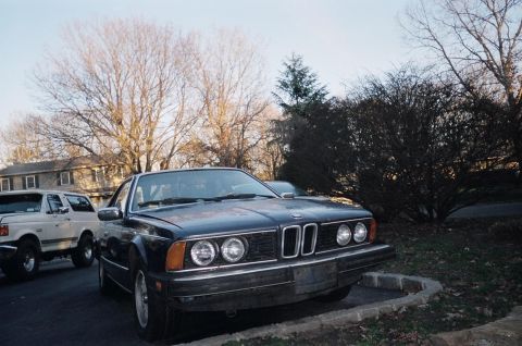 1983 BMW 5 Series - A dark blue BMW coupe is parked on the street next to a white SUV, with bare trees and houses visible.