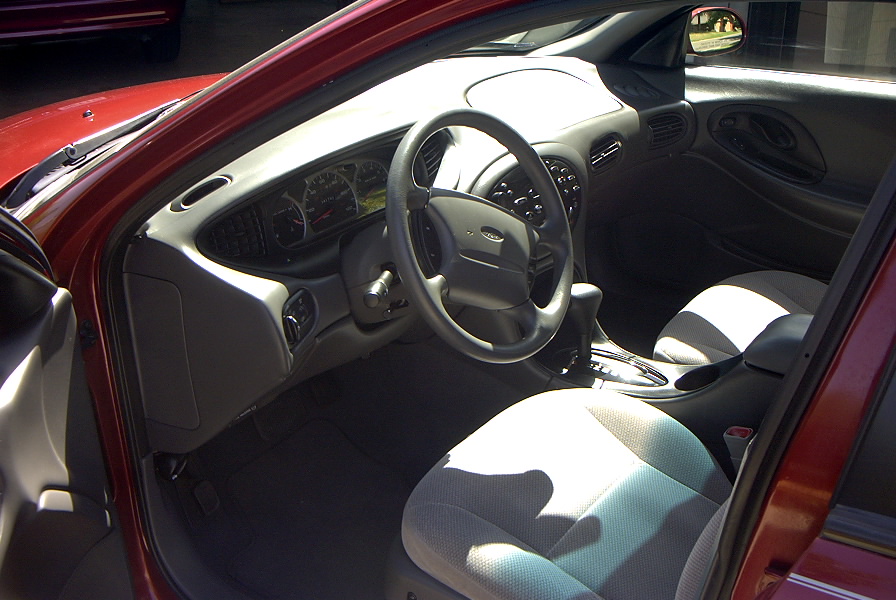 Red car interior with gray dashboard, steering wheel, and cloth seats. "SUPERMOTORS" text visible