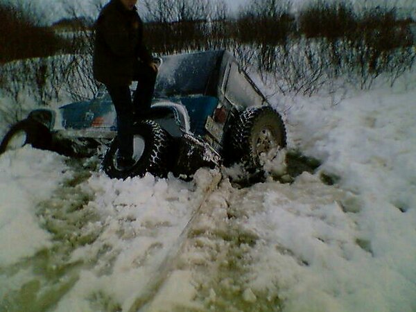 Blue off-road vehicle with large tires stuck in deep snow, a person stands on the hood