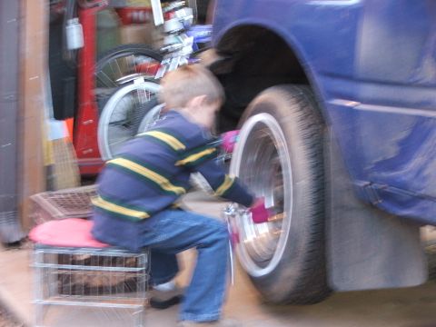 1990 Dodge Shadow - A young boy sits on a makeshift stool to work on the wheel of a blue car, which appears to have custom rims.