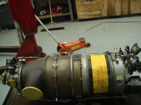 1996 Ford Bronco - A jet engine sits on a workshop floor, supported by a red stand and an orange jack, with a caution label visible.