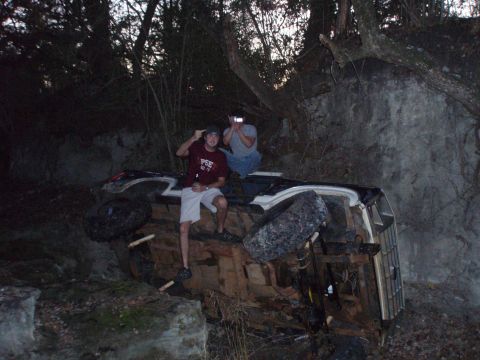 1986 Ford Bronco - Two individuals sit atop an overturned 1986 Ford Bronco, resting against a rocky embankment.