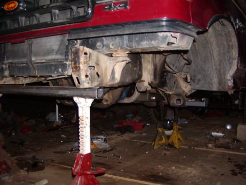 1991 Ford Bronco - Front end of a red 1991 Ford Bronco, elevated on jack stands for repair, revealing the chassis and suspension components.