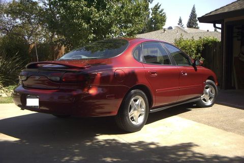 1999 Ford Taurus - The rear of a red sedan is visible, parked next to a garage with trees in the background.