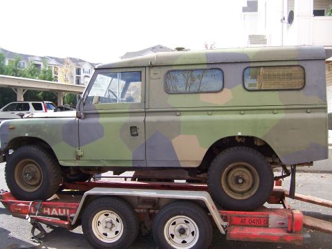 1974 Land Rover Land Rover - This camouflaged Land Rover Defender is being transported on a trailer, showing its side profile with a hardtop enclosure.
