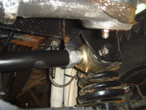 1996 Ford Bronco - Underbody close-up of a black tie rod or drag link threaded into a brass-colored heim/rod-end joint bolted through a steel bracket, with a…