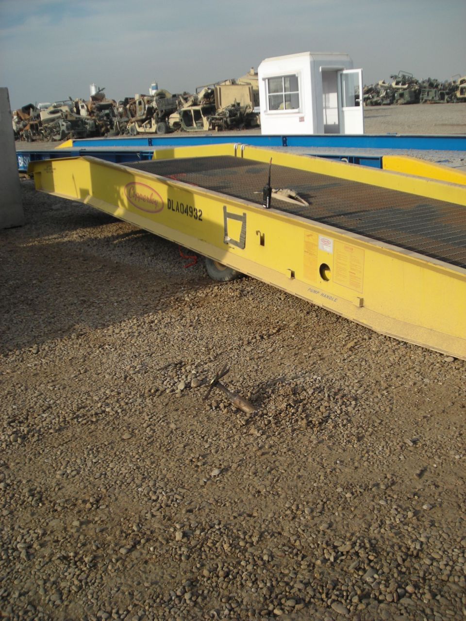 Yellow trailer with a radio on it, parked on gravel. Military vehicles are in the background
