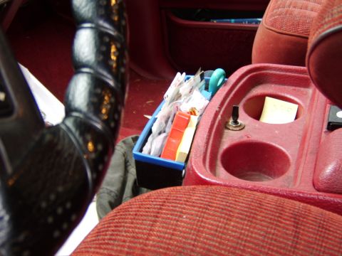 1989 Ford Bronco - The interior console of a vehicle, cup holders and an organizer filled with various packets and items.