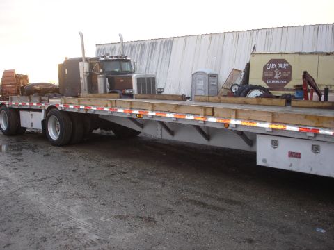 2006 Ford F-350 - A flatbed trailer carries a semi-truck, a portable toilet, and other equipment, with a building in the background.