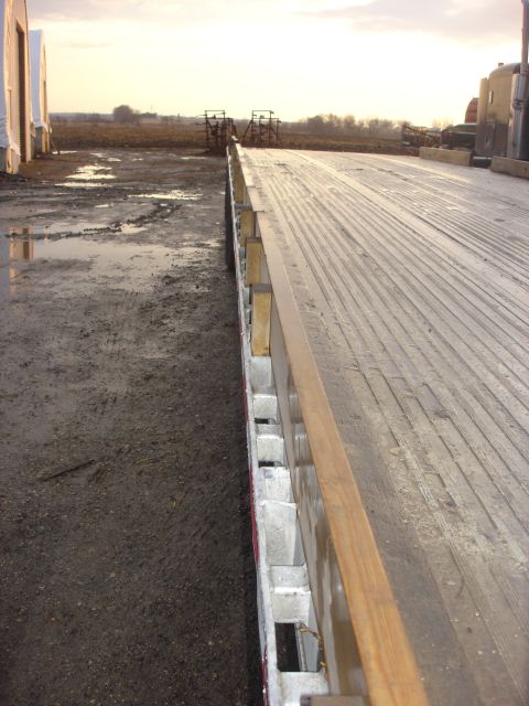2006 Ford F-350 - A view of a flatbed trailer's wooden deck next to a muddy area with some structures in the distance.