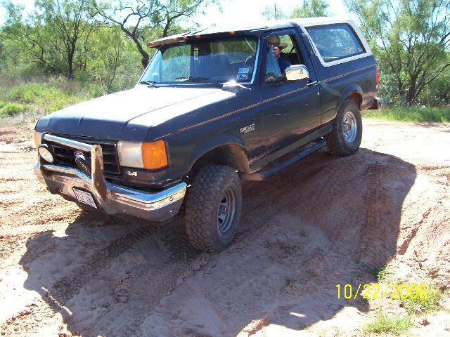 Dark green 1987 Ford Bronco with white top driving on a dirt hill. Bull bar on front
