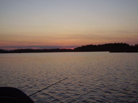 1996 Ford F-150 - A fishing rod extends from a boat into a calm lake as the sun sets over a distant tree line.