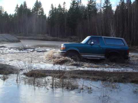 1996 Ford Bronco - A teal Ford Bronco navigates a treacherous path of mud and ice, kicking up water as it goes.
