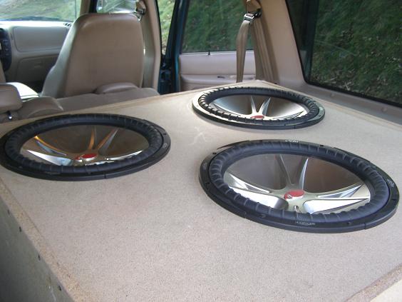 Three subwoofers in a tan carpeted enclosure filling an SUV cargo area