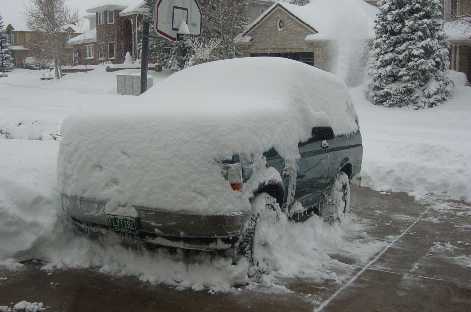 Dark green SUV buried in deep snow in a driveway, Colorado plate reading FLYTIRE visible on front bumper