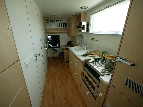 Interior of a vehicle's kitchen area with light wood cabinets, a sink, a stove, and a window