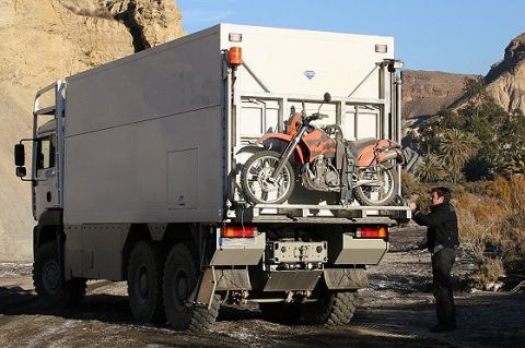 1993 Ford Bronco - A man secures a dirt bike onto the back of a large, multi-wheeled truck in a rocky, desert environment.
