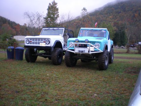1969 Ford Bronco - Two modified Ford Broncos, one white and one blue, are parked on a grassy area with a wooded hillside behind them.
