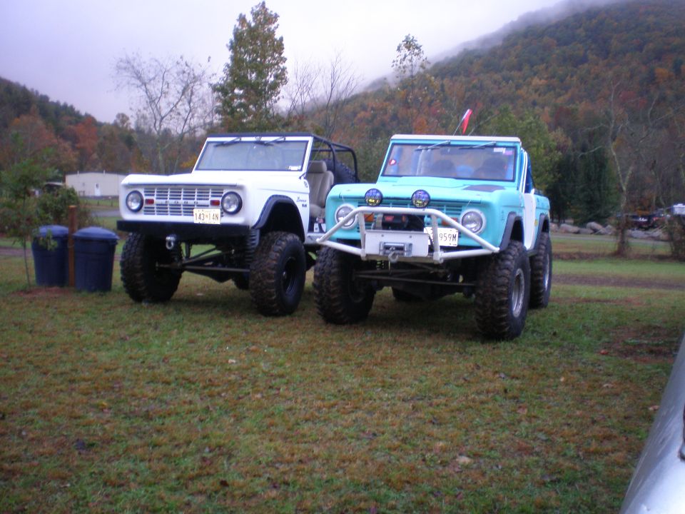 1969 Ford Bronco and a blue Ford Bronco parked side-by-side on grass with trees and hills in the background