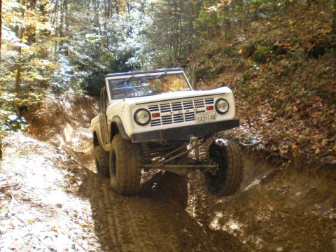1969 Ford Bronco - A white 1969 Ford Bronco navigates a steep, muddy off-road trail surrounded by trees.