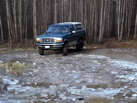 1996 Ford Bronco - A teal Ford Bronco with a white roof is parked on a dirt road in a wooded area.