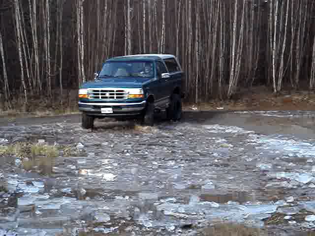 1996 Ford Bronco driving on a dirt road with ice patches, surrounded by trees