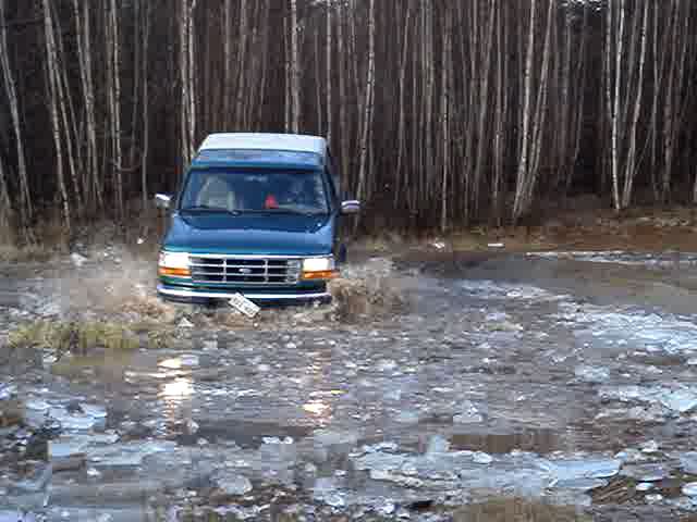 Teal Ford Econoline van driving through icy water, front view