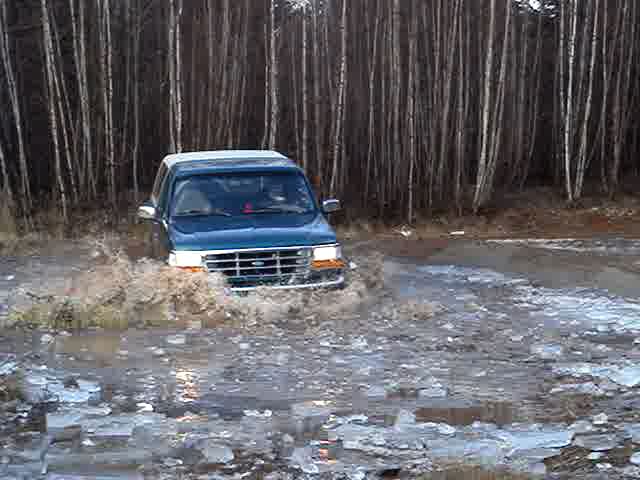 Blue Ford Bronco driving through a muddy, icy puddle with trees in the background