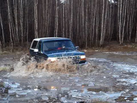 1996 Ford Bronco - A 1996 Ford Bronco splashes through water and mud near a wooded area.