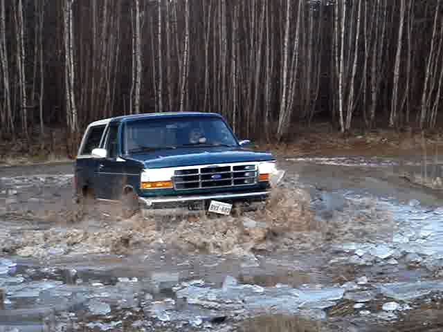 Dark blue Ford Bronco driving through muddy water with ice chunks. Front grille and headlights visible