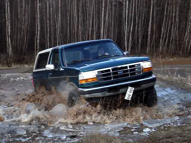 1996 Ford Bronco driving through muddy water with trees in background