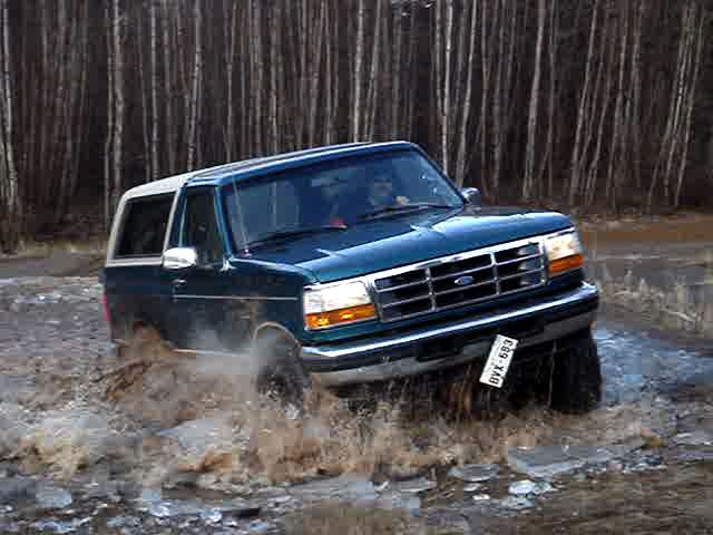 Blue 1996 Ford Bronco driving through muddy water with trees in background