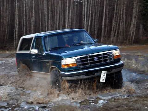 1996 Ford Bronco - A Ford Bronco navigates a muddy terrain, kicking up water as it drives.