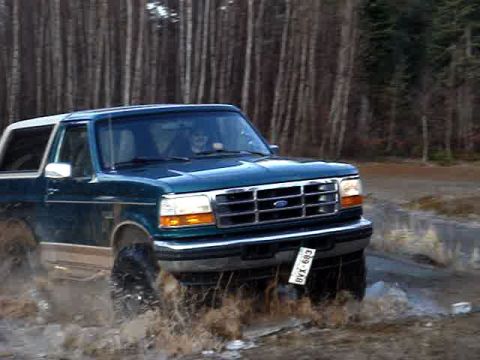 1996 Ford Bronco - 1996 Ford Bronco splashes through water on a dirt road, surrounded by a forest.