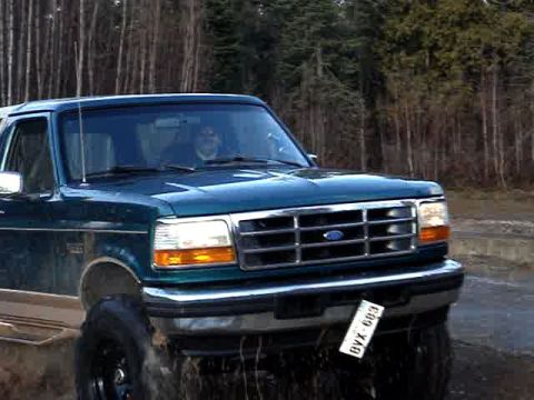 1996 Ford Bronco - A teal Ford Bronco is shown from the front-right, driving on a dirt road with trees in the background.