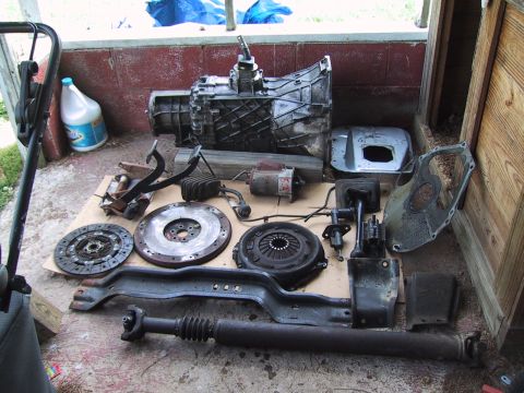 1994 Ford Bronco - A complete manual-transmission swap kit laid out on cardboard in a shed: a manual gearbox with shifter, bellhousing, flywheel, clutch disc…