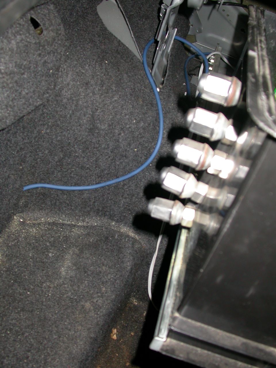 Car trunk interior with gray carpet, blue wire, and a row of lug nuts attached to a metal bracket