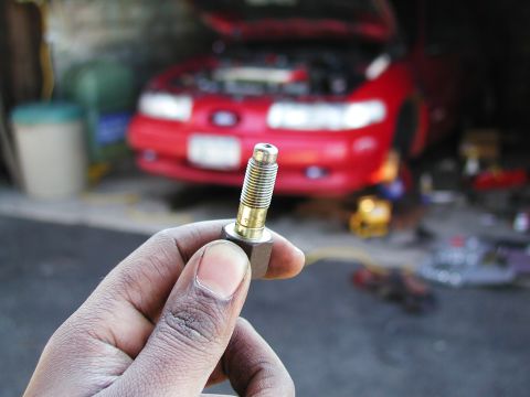 1992 Ford Taurus - A hand holds a small threaded brass/steel fitting with a hex flange up to the camera, likely a bleeder or sensor-style screw. Behind it, ou…