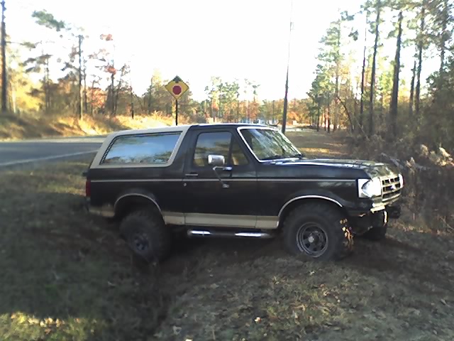 Black and tan Ford Bronco with lifted tires parked on a grassy roadside slope, side view