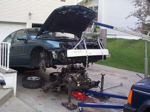 1997 Ford Taurus - A teal/green four-door sedan sits in a driveway with its hood up and front subframe and engine assembly lowered out onto a floor jack and e…