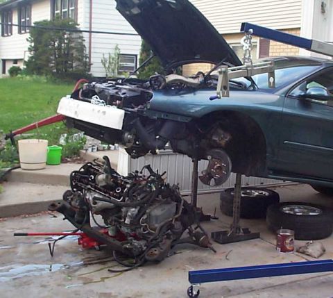1997 Ford Taurus - A green sedan sits nose-up on jack stands with its hood open and engine bay emptied, while the removed engine and transaxle assembly rests…