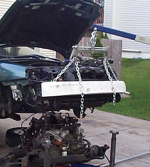 1997 Ford Taurus - An engine hoist with a blue boom and chains slung under a white load bar lifts the front of a teal sedan with its black-lined hood raised a…