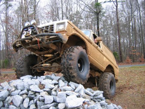 1992 Ford Mustang - A lifted, mud-covered SUV navigates a pile of rocks on a wooded trail.