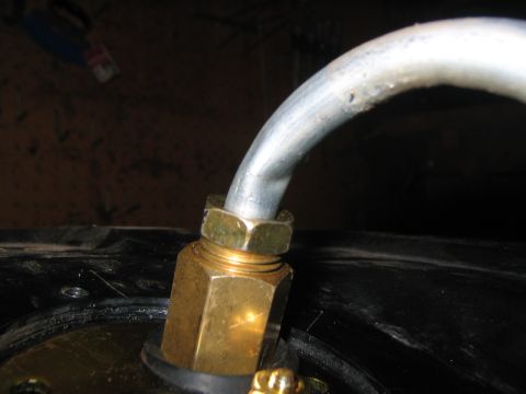1979 Ford Bronco - Close-up of a brass compression fitting threaded into the sending-unit flange on top of a black fuel tank, with a bent steel fuel line curv…