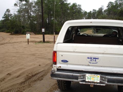 1989 Ford Bronco - A white Ford Bronco is parked on a sandy track, with trees and a signpost visible.